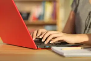 Single student hands writing on line in a red laptop sitting in a table with a notebook beside at home with a homey background