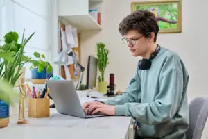 Young male college student sitting at desk at home using laptop. Handsome guy 19 years old with glasses typing on keyboard. Use internet online technologies for study, leisure, communication