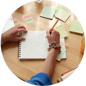 Close up of a studewnt taking notes at a desk, surrounded by Post-its, with a glass of orange juice at his side.
