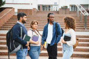 Diverse group of young students with books and backpacks interacting and discussing outside a university building
